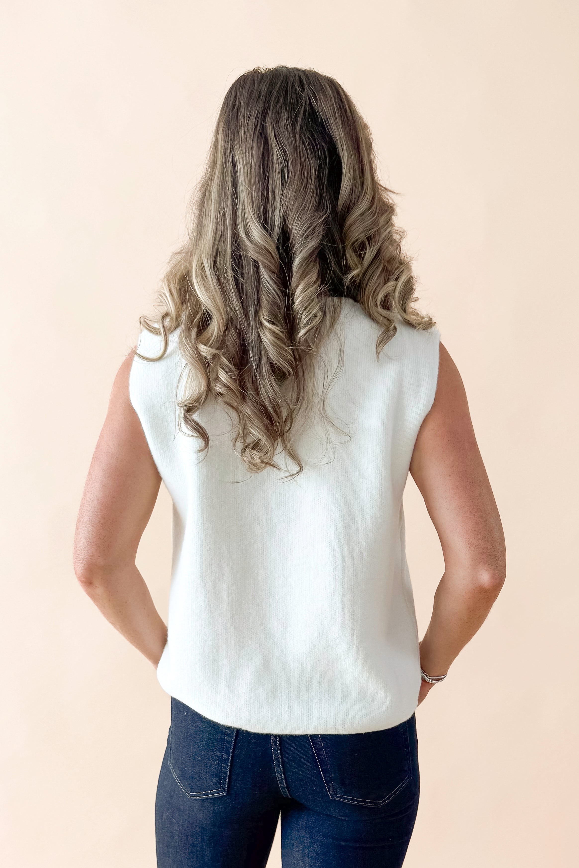 Back view of woman wearing Cream Button Detail Sweater Vest with jeans against tan studio background.