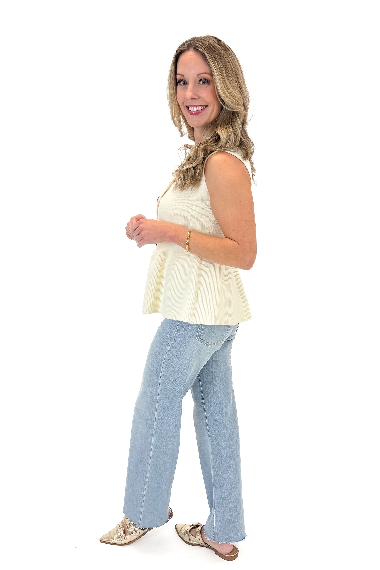 Side view of woman wearing Cream Peplum Sweater Tank with jeans against white studio background.