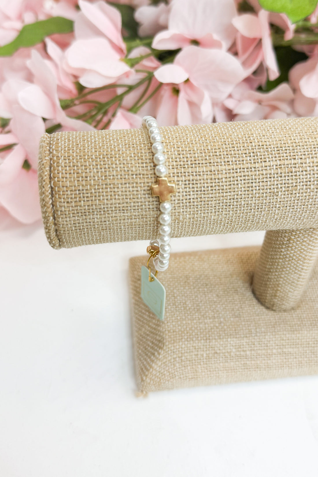 Pearl bracelet with a gold cross and tag on a beige jewelry stand with pink flowers in the background