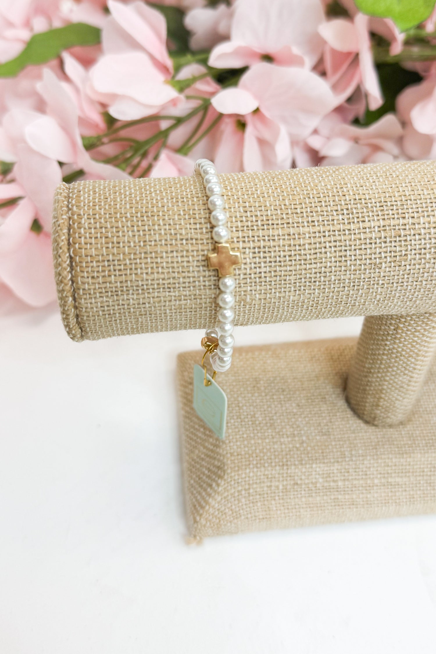 Pearl bracelet with a gold cross and tag on a beige jewelry stand with pink flowers in the background