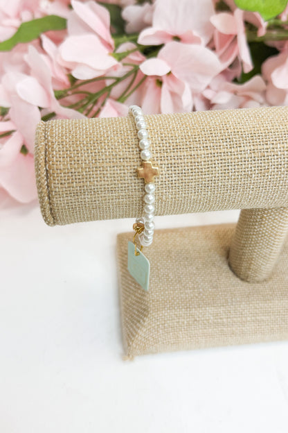 Pearl bracelet with a gold cross and tag on a beige jewelry stand with pink flowers in the background