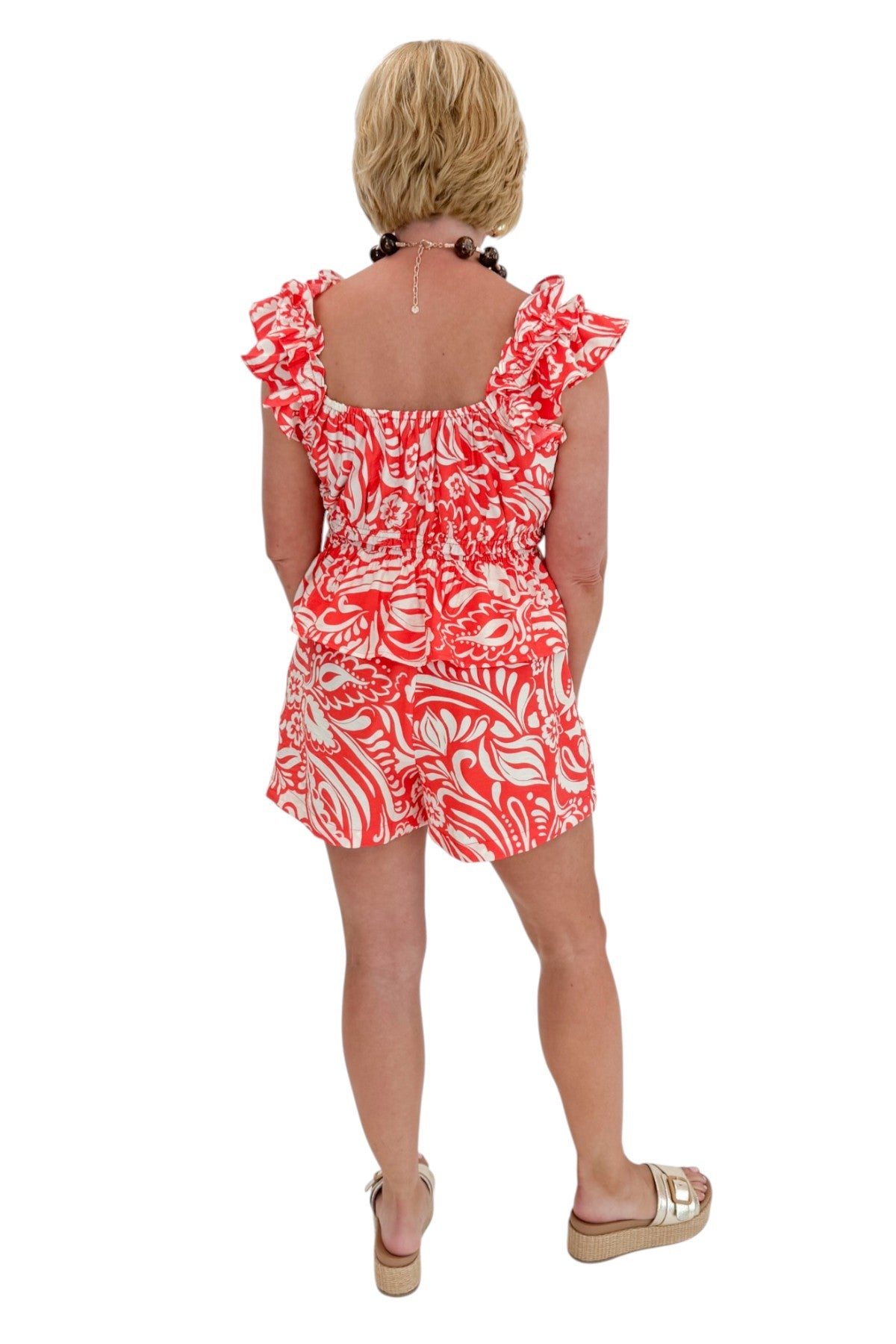 Back view of woman wearing Swagger Boutique’s Coral Printed Smocked Peplum Tank with matching Coral Printed Shorts against white studio background.