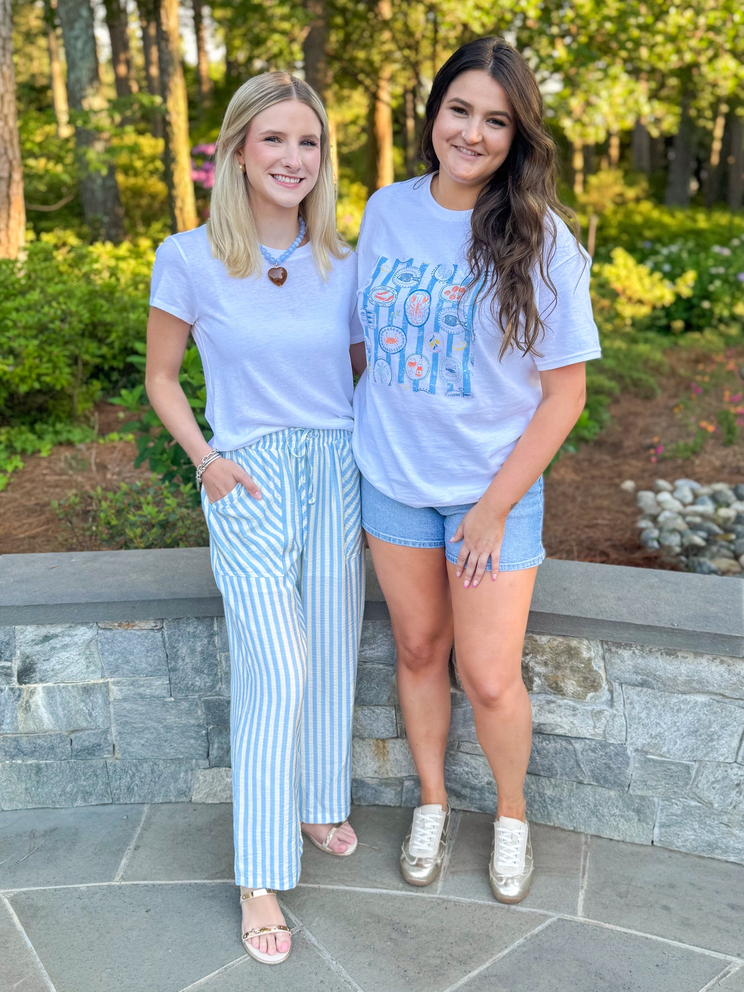 Front view of woman wearing Swagger Boutique’s Blue &amp; Cream Striped Pants with white top standing outside with woman wearing tee and jean shorts.