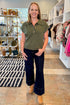 Woman in a green shirt and dark jeans standing in a store with shelves and clothing in the background.