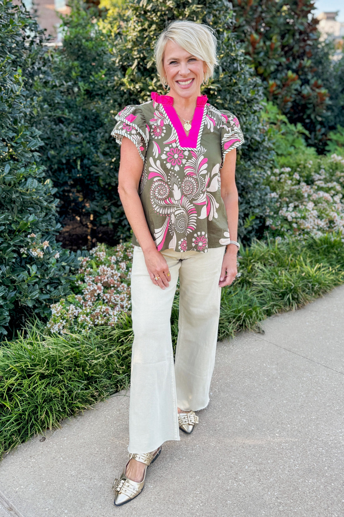 Woman in a patterned top and white pants standing outdoors with greenery in the background