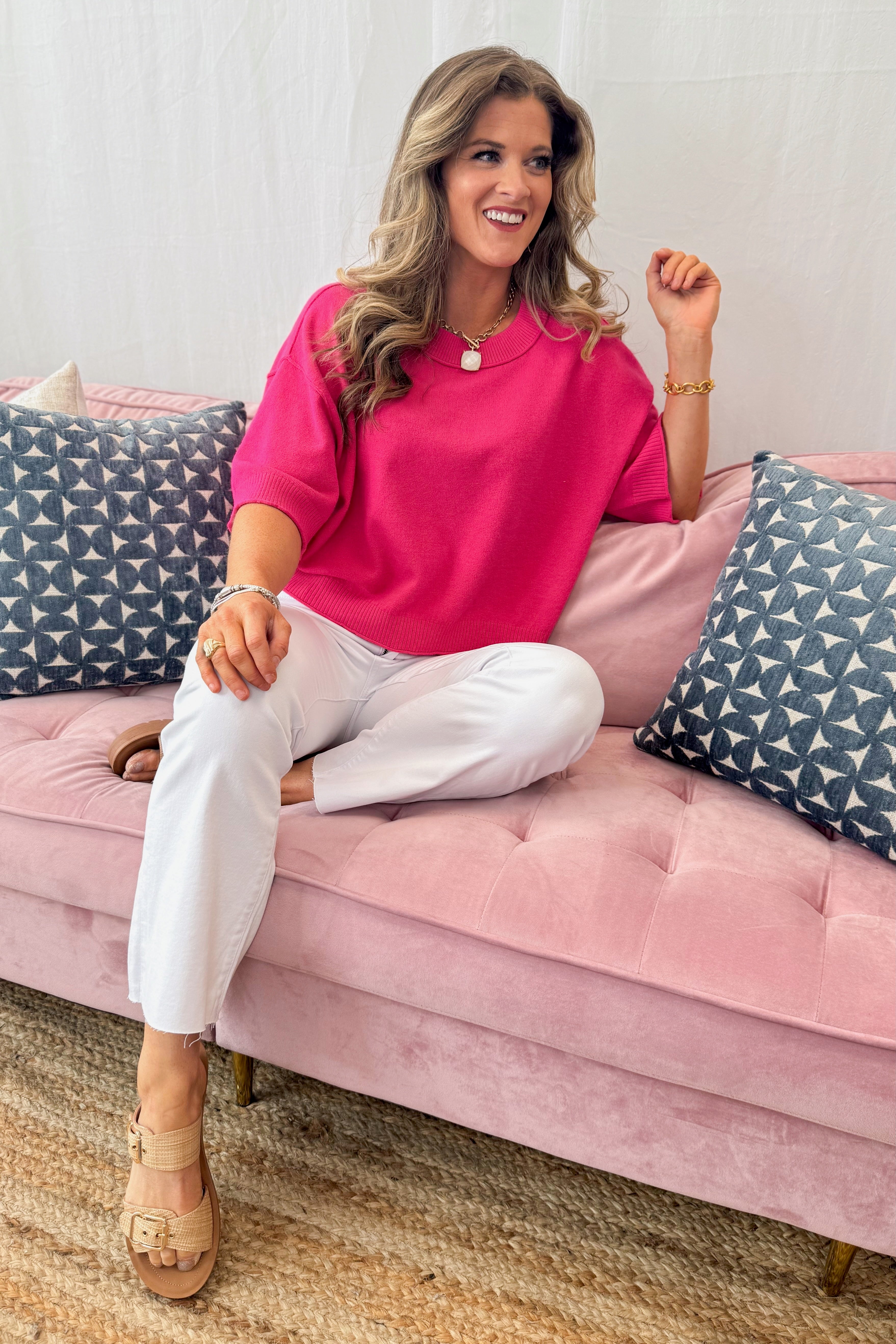 Woman in a pink shirt and white pants sitting on a pink couch with patterned cushions.
