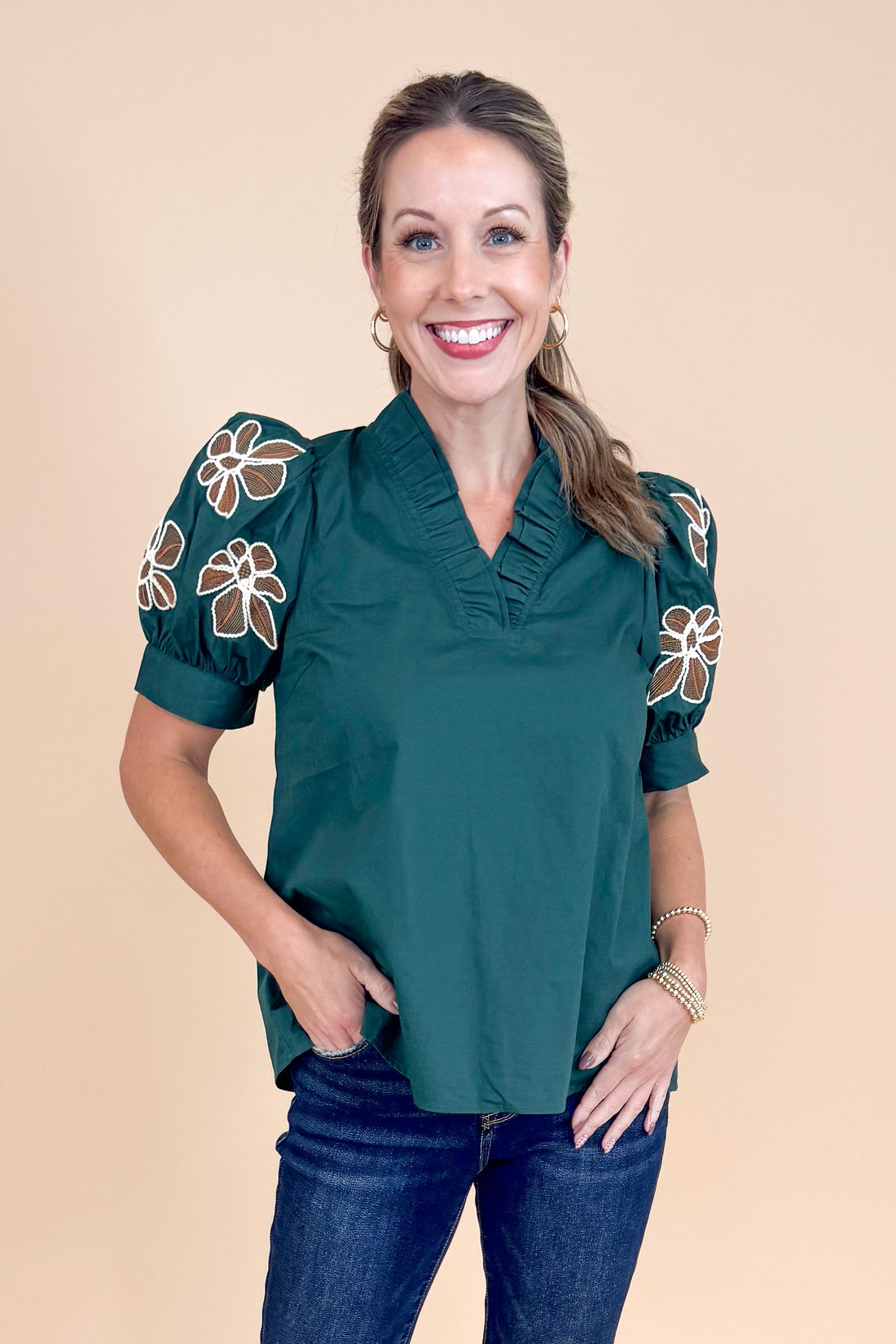 Woman wearing a dark green blouse with floral patterns on sleeves against a beige background