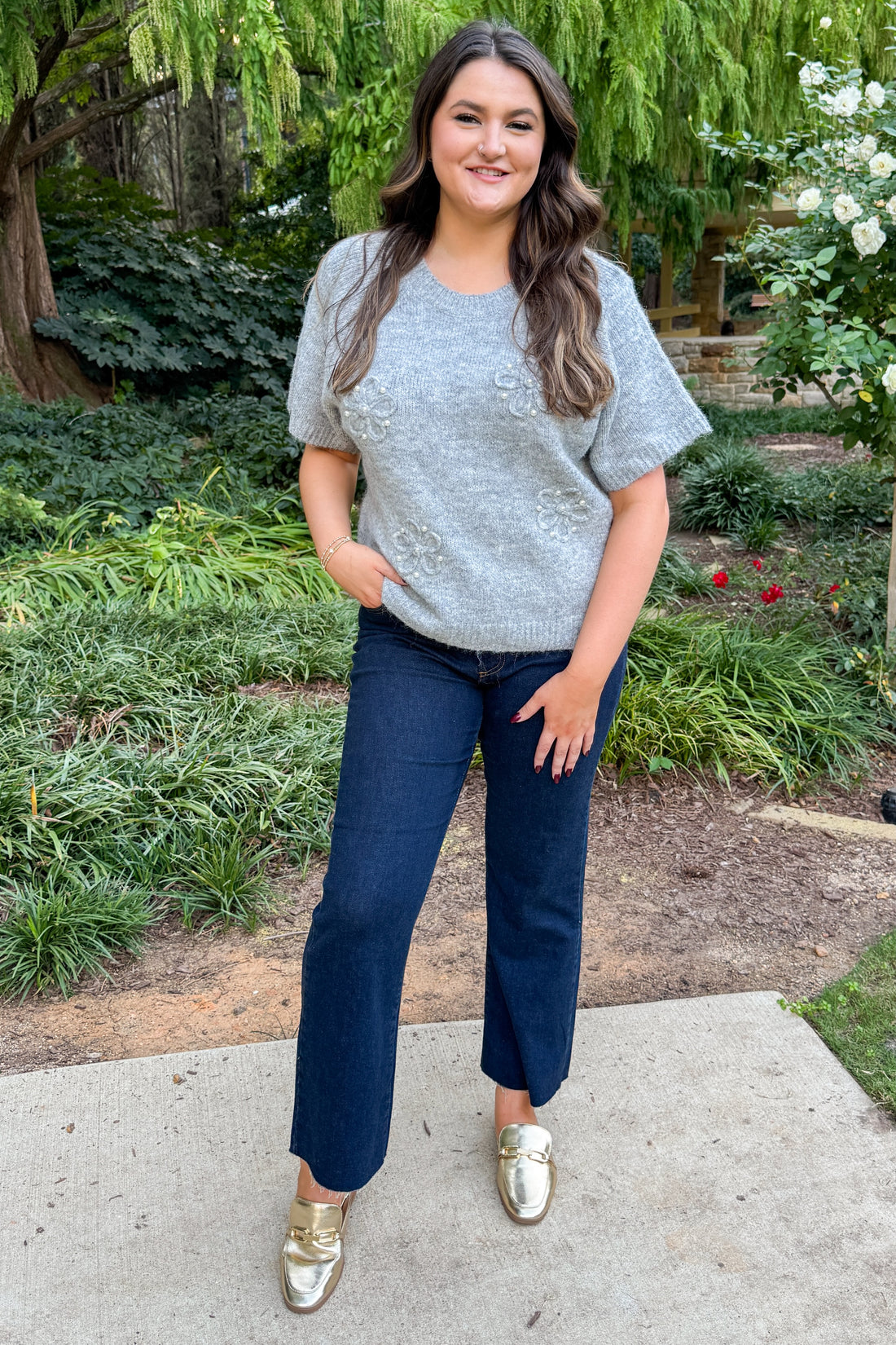 Woman wearing Grey Floral & Pearl Detail Short Sleeve Sweater by Entro and blue jeans while outside with greenery around.