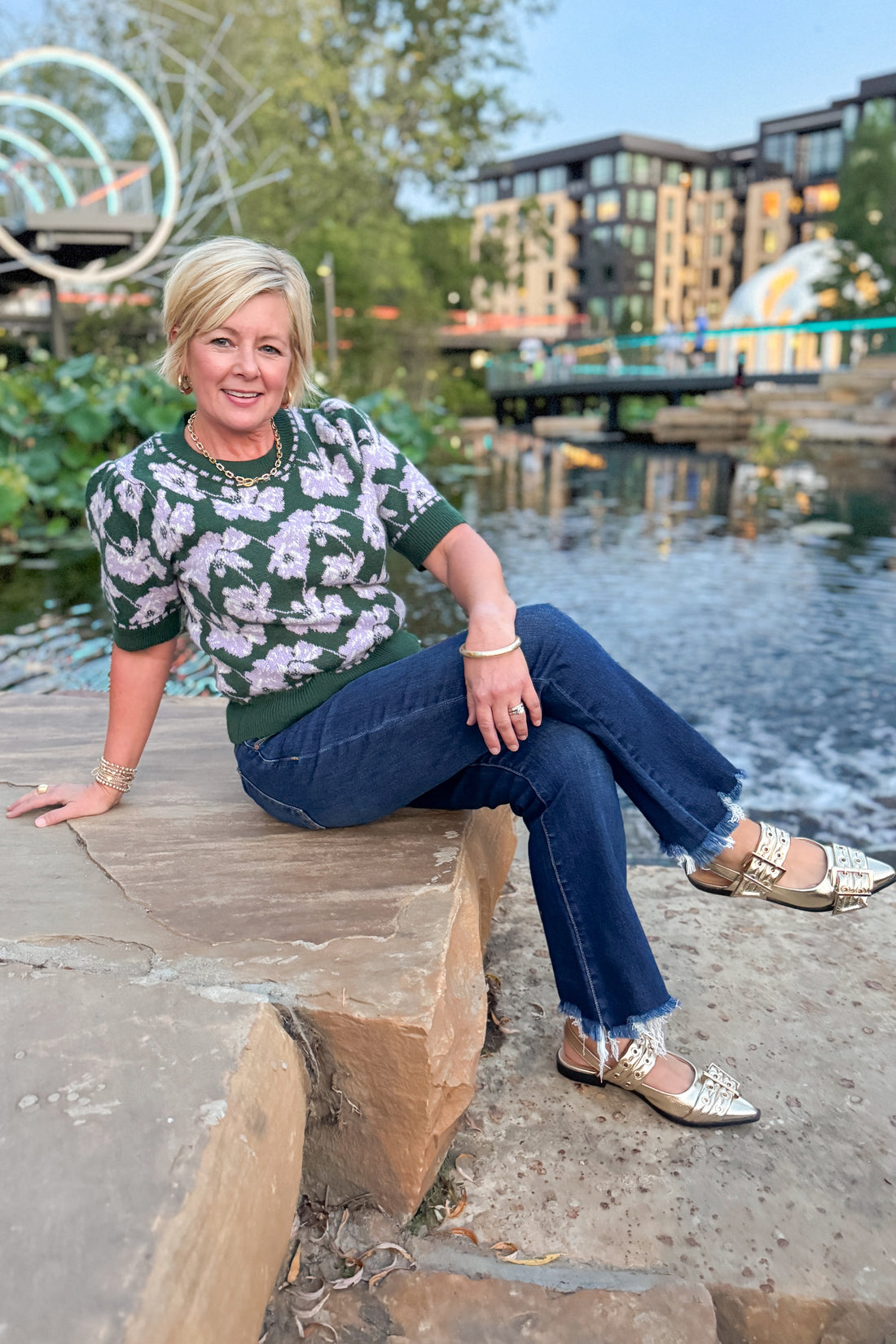 Woman sitting on a stone ledge by a waterfront with buildings and greenery in the background