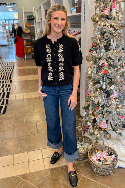 Front view of woman wearing Black Center Embroidered Short Sleeve Cardigan with jeans against holiday decor.