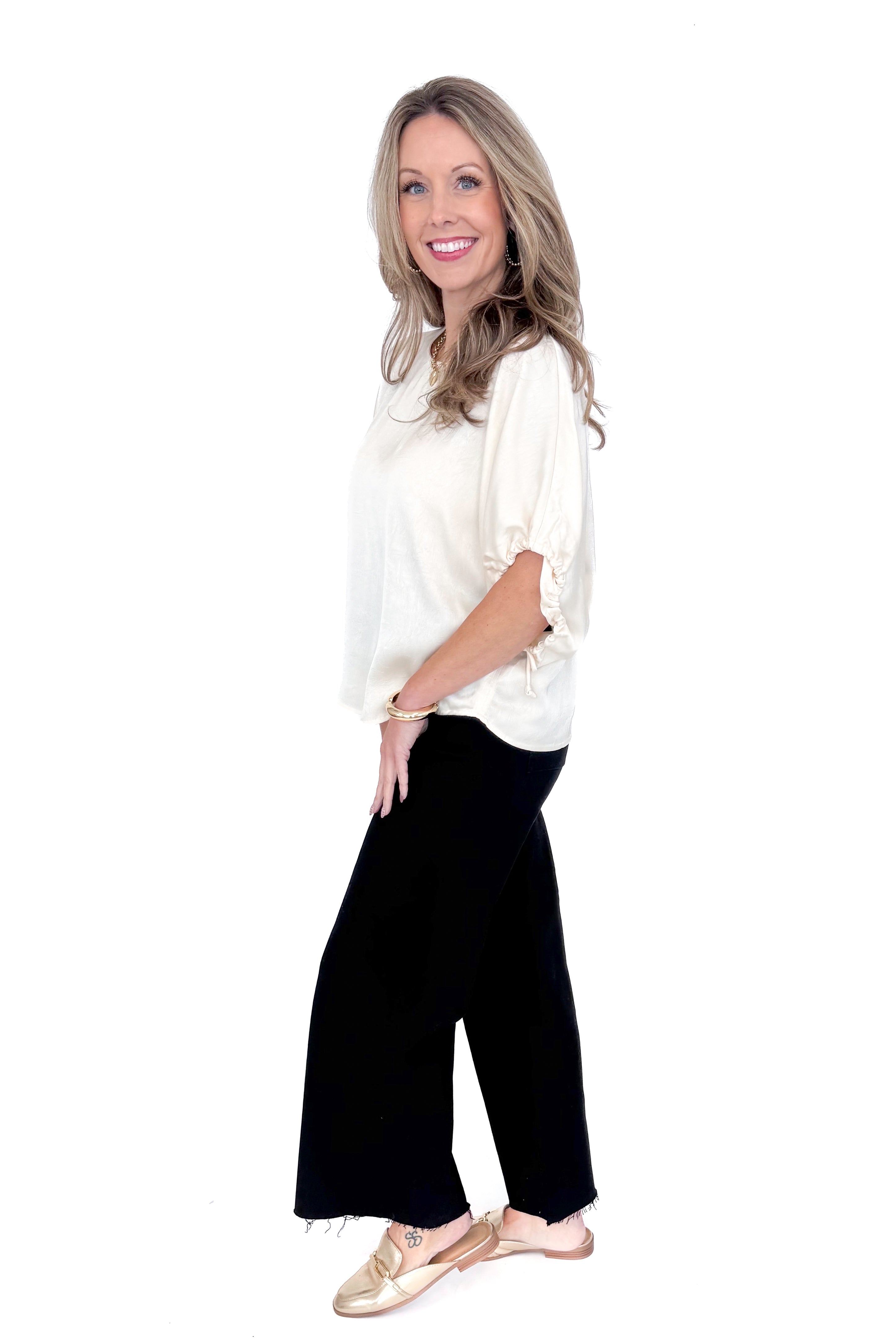 Side view of woman wearing Champagne Satin Tie Sleeve Top with jeans against white studio background.