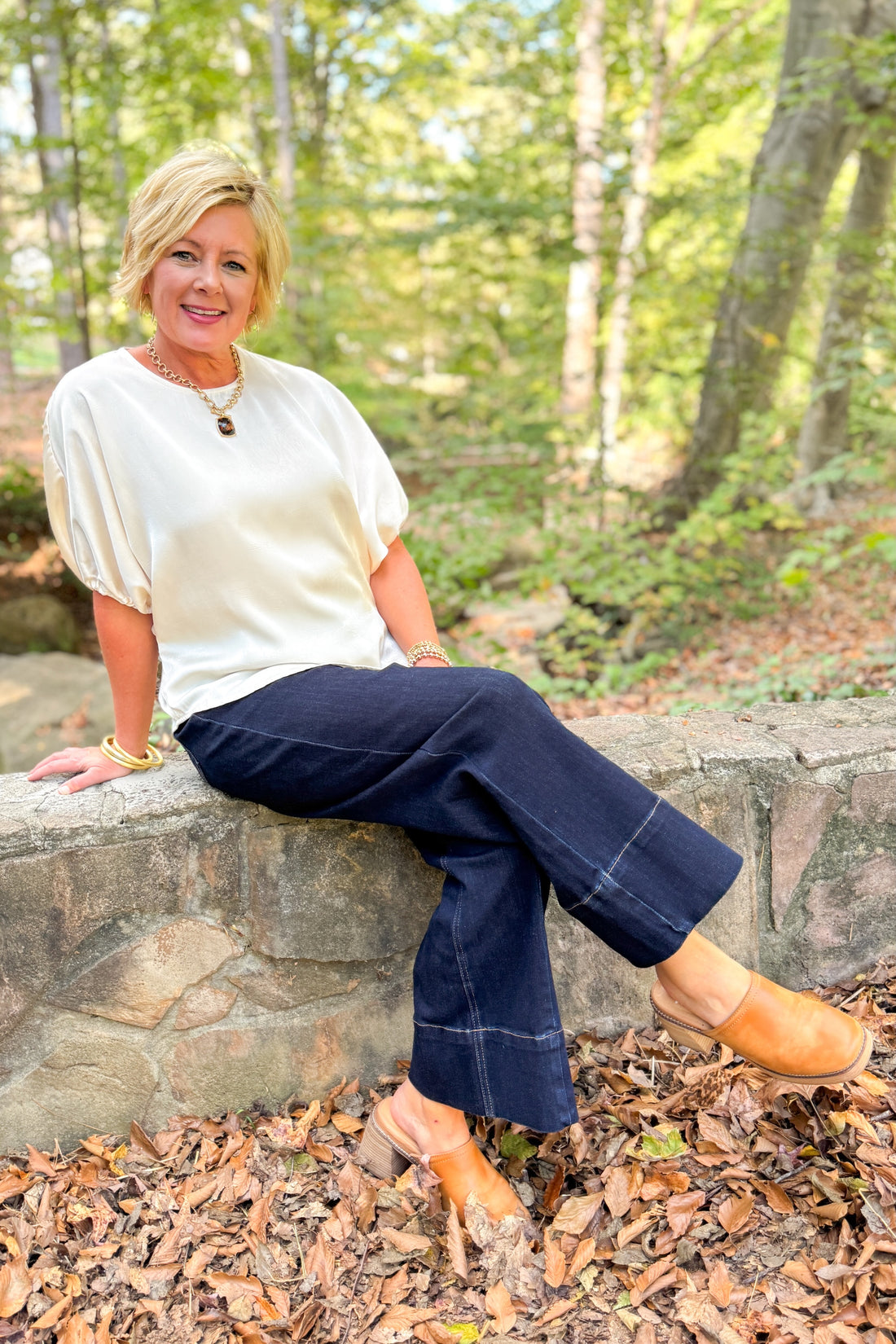Front view of woman sitting outside wearing Champagne Satin Tie Sleeve Top with jeans with leaves and trees around.