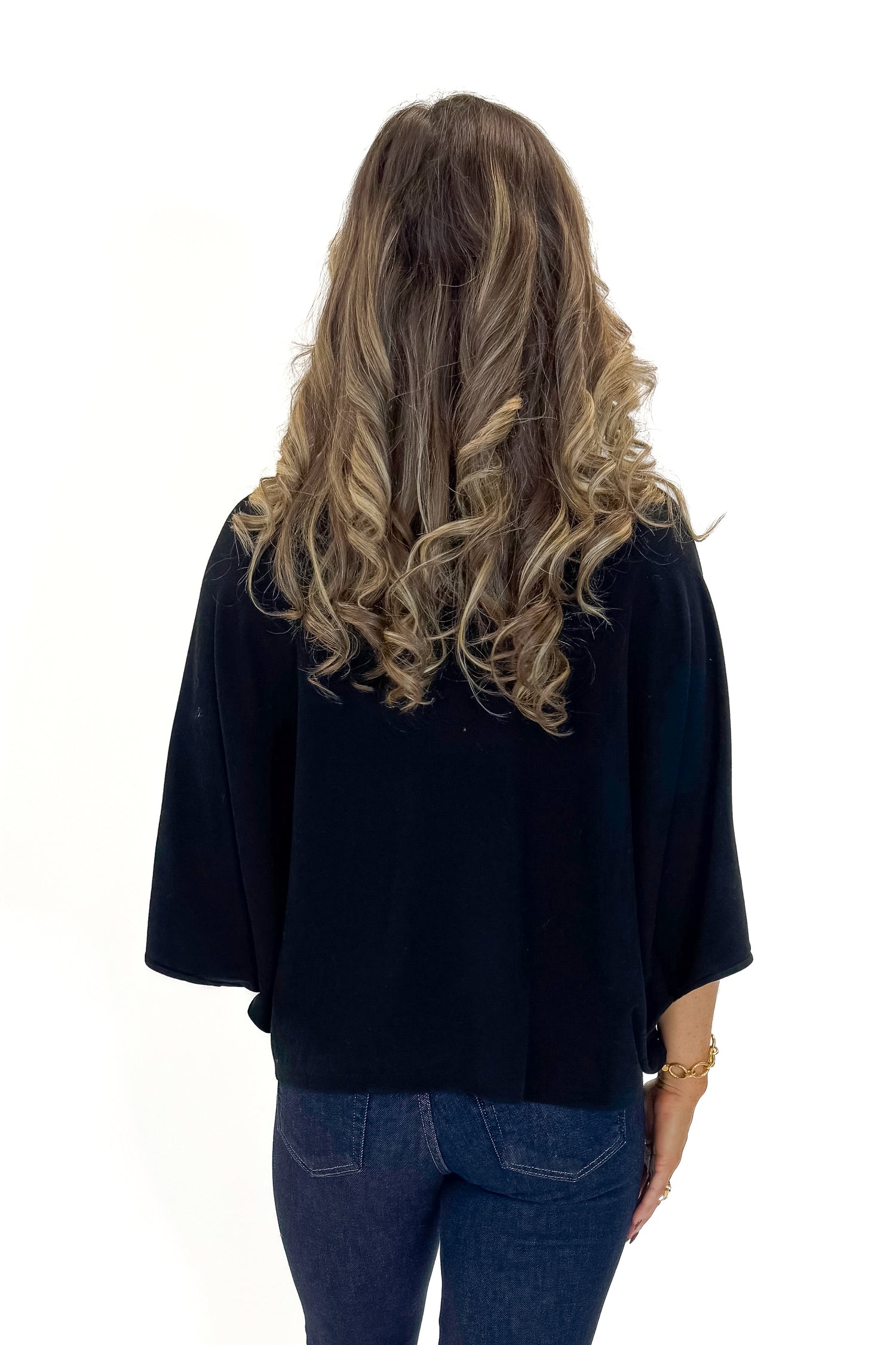 Back view of woman wearing Black Mock Neck Poncho Sweater with jeans against white studio background.