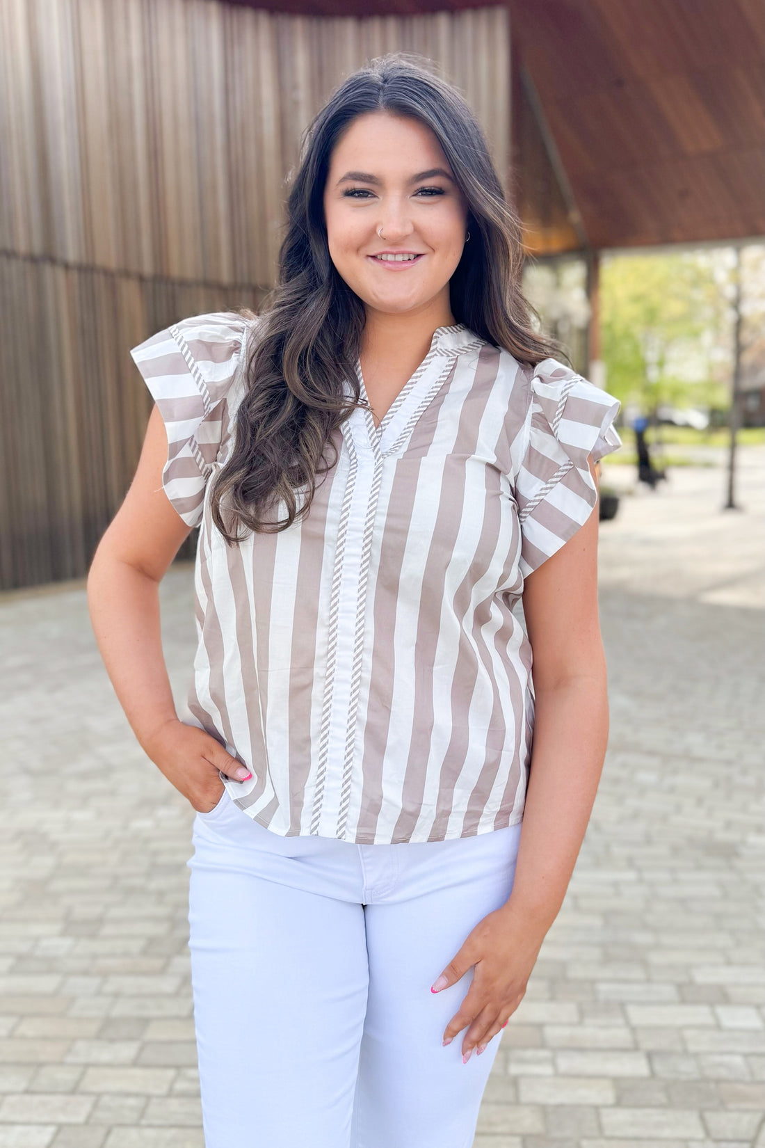 Front view of woman wearing Taupe Striped Flutter Sleeve Top by Entro with jeans outside at a park.