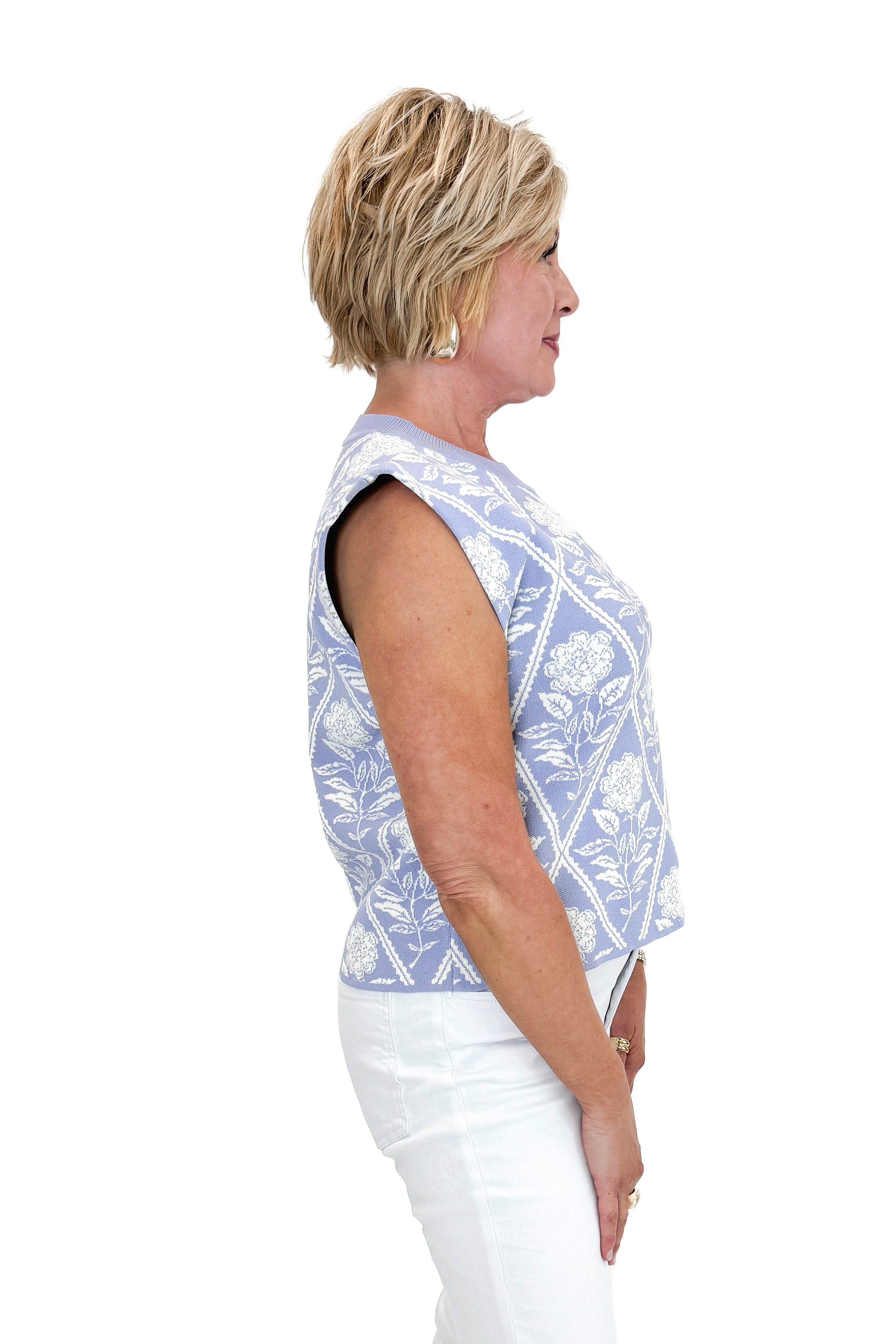 Side view of woman wearing Periwinkle Floral Printed Sleeveless Sweater by Entro with white jeans against white studio background.