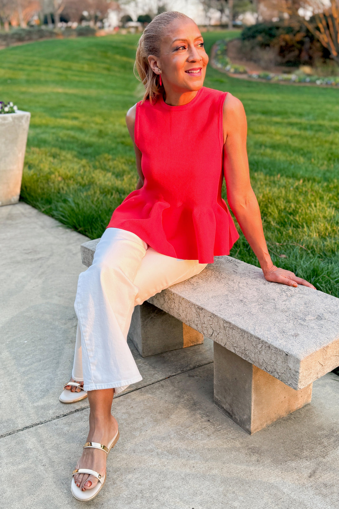 Front view of woman wearing Red Peplum Sweater Tank by Entro with jeans outside sitting on a bench.
