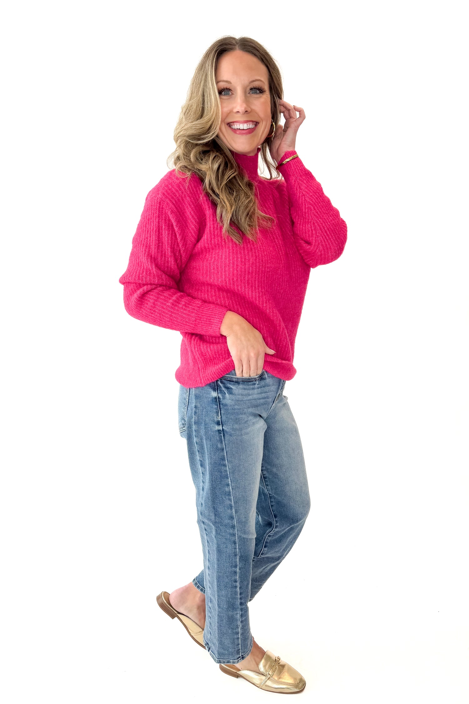Side view of woman wearing Hot Pink Ribbed Turtleneck Sweater with jeans against white studio background.