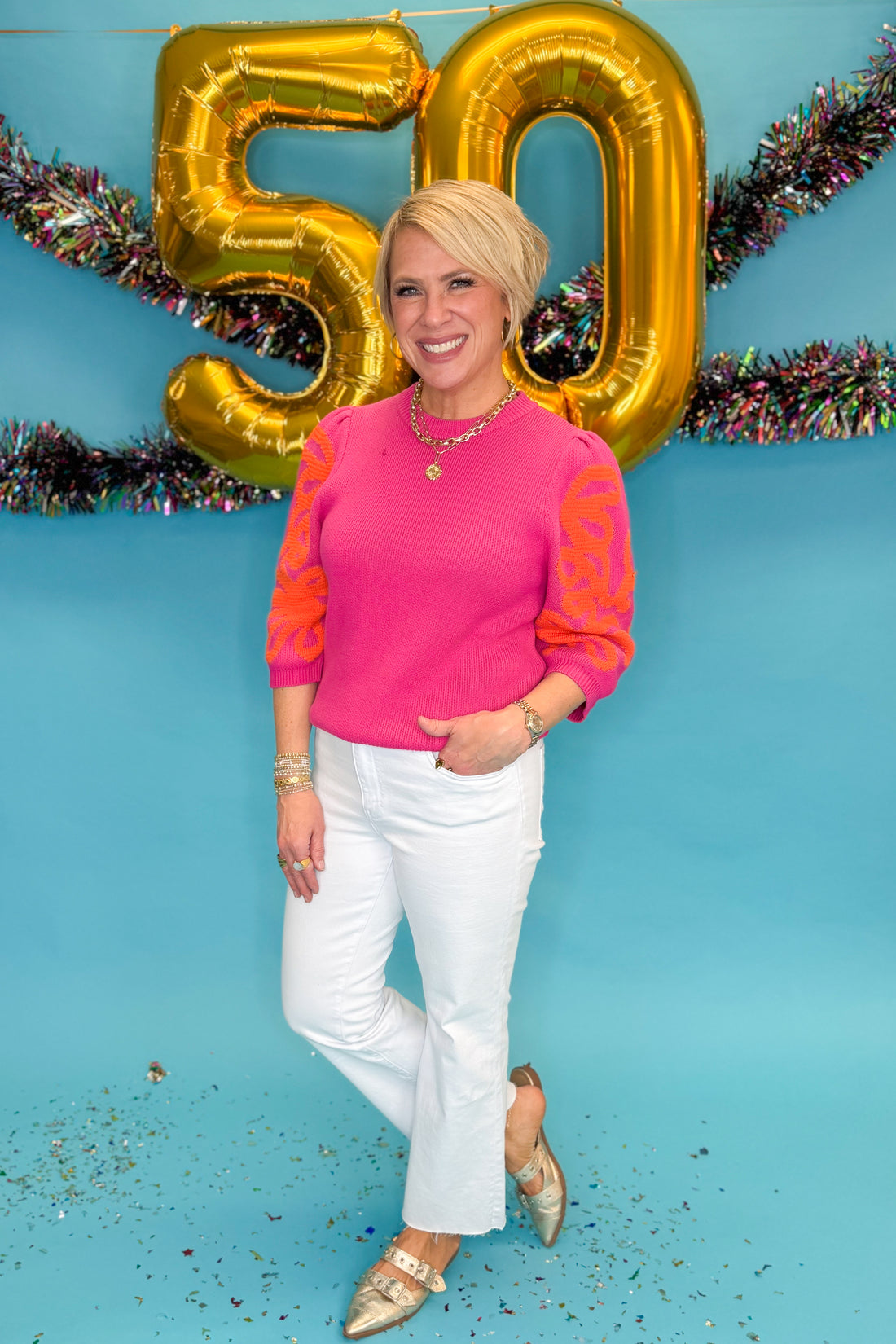 Front view of woman wearing Pink &amp; Orange Embroidered Short Sleeve Sweater with jeans against blue background.