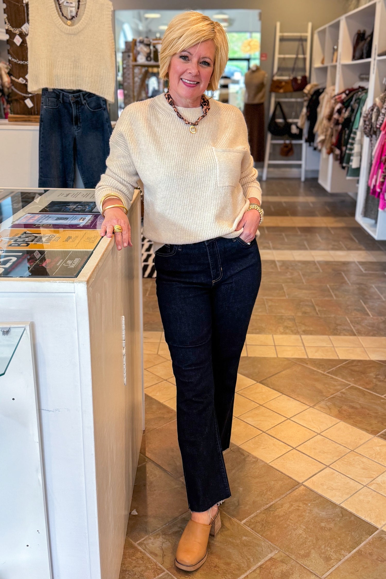 Woman in a store wearing a white sweater and dark jeans, standing next to a display case.