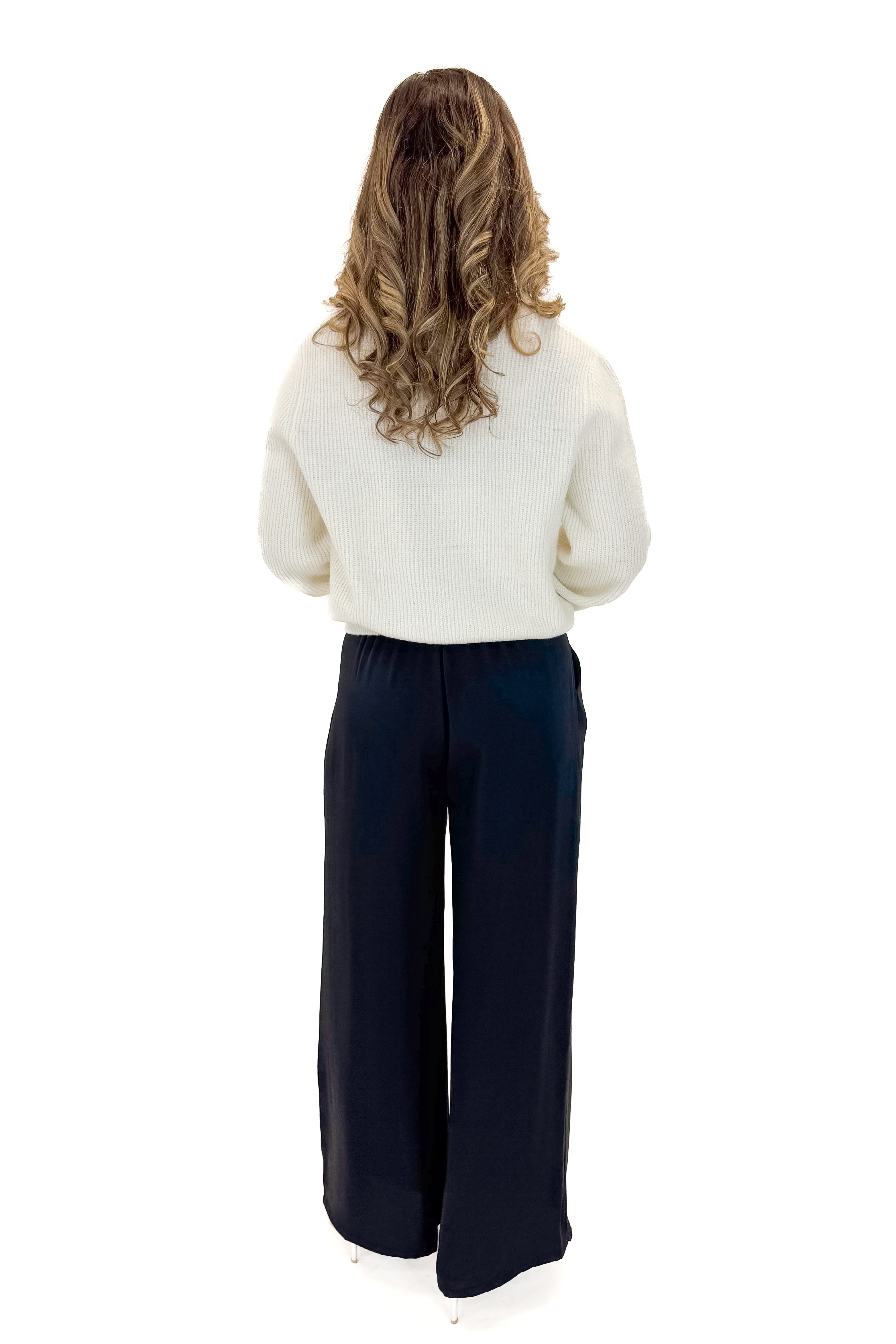 Back view of woman wearing sweater with Black Satin Pants against white studio background.