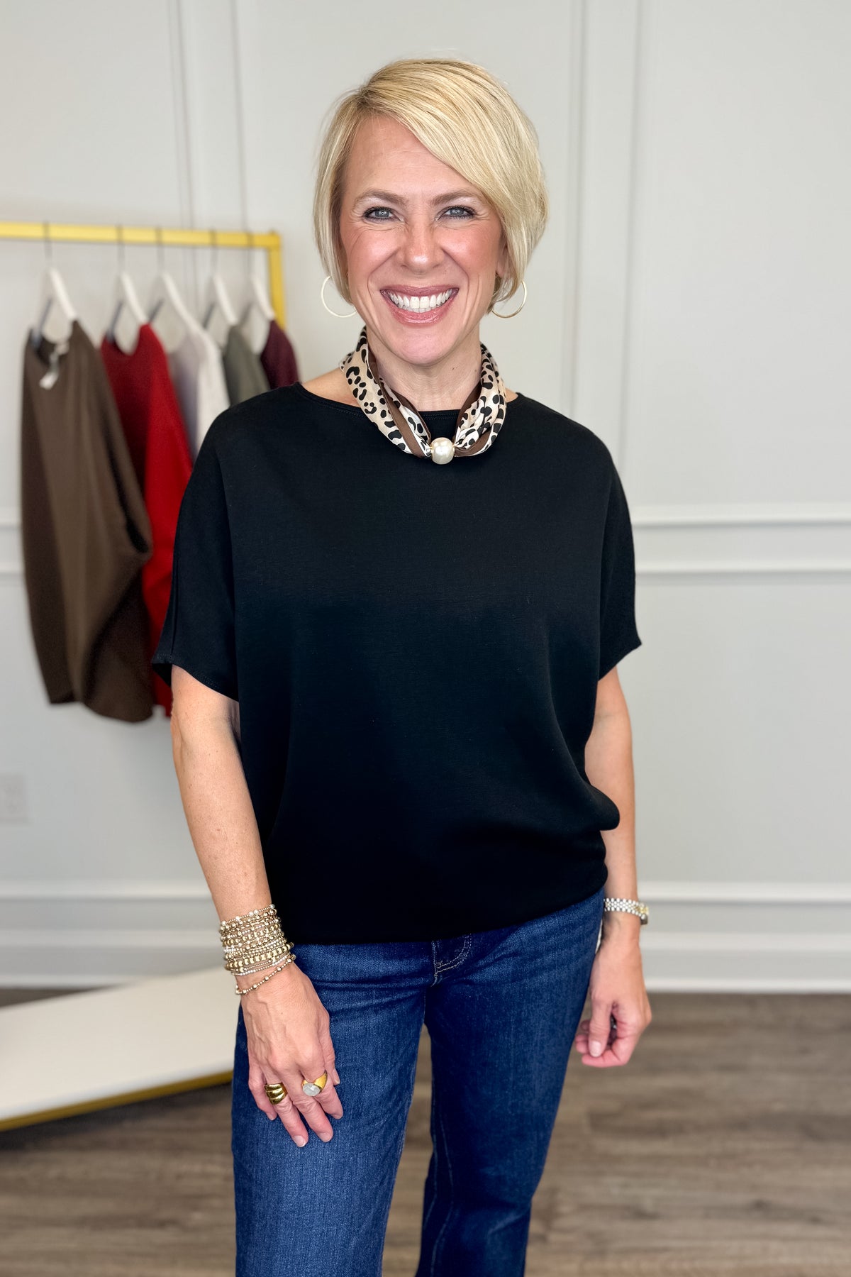 A smiling blonde woman stands indoors wearing a loose black short-sleeve top, blue jeans, layered statement necklaces, bracelets, and rings. A clothing rack with other garments is visible in the background against a light wall.