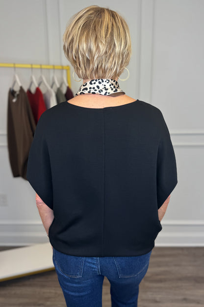 A woman viewed from behind wearing a black short-sleeve top and blue jeans, standing in a boutique setting with a clothing rack visible in the background.