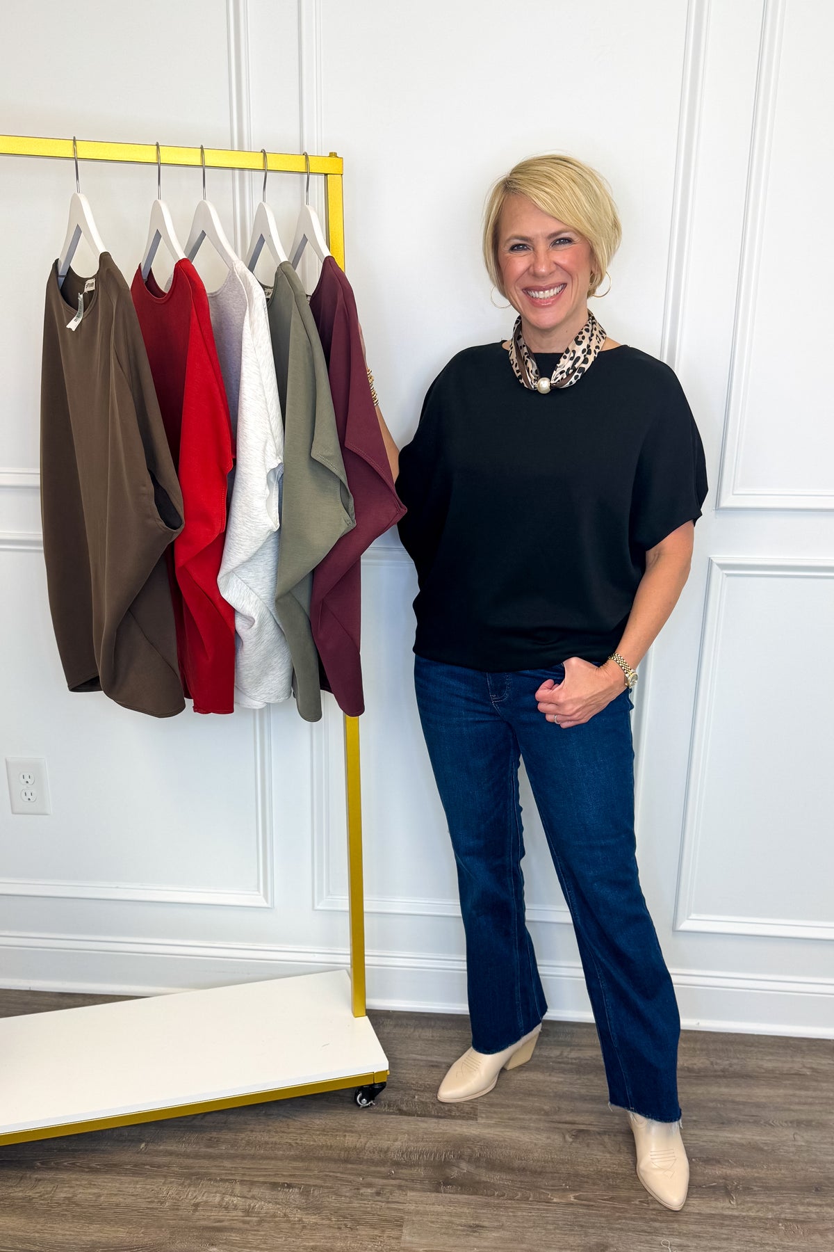 A woman with short blonde hair stands next to a yellow clothing rack in a room with white paneled walls and wood floors. She is wearing a black short-sleeve top, dark blue jeans, and beige ankle boots.