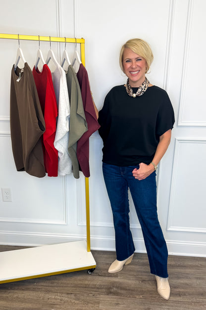 A woman with short blonde hair stands next to a yellow clothing rack in a room with white paneled walls and wood floors. She is wearing a black short-sleeve top, dark blue jeans, and beige ankle boots.