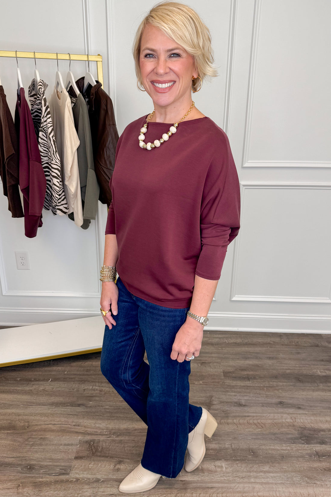 A woman modeling a burgundy long-sleeve knit top with dark blue jeans and cream ankle boots, standing in a boutique setting with clothing racks and paneled walls in the background.