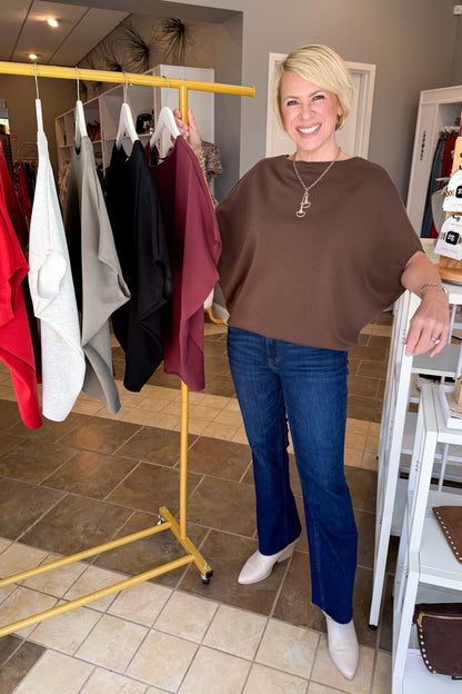 A smiling blonde woman stands in a boutique wearing a brown short-sleeve top, blue jeans, and white ankle boots. A yellow clothing rack with several garments is visible behind her, along with store fixtures and tiled flooring.
