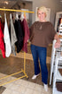 A smiling blonde woman stands in a boutique wearing a brown short-sleeve top, blue jeans, and white ankle boots. A yellow clothing rack with several garments is visible behind her, along with store fixtures and tiled flooring.