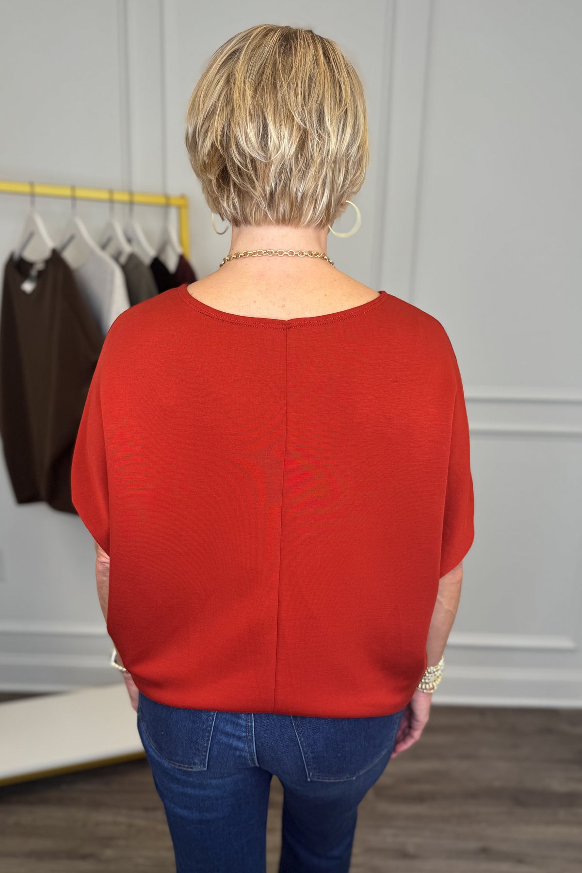 A woman viewed from behind wearing a bright red short-sleeve knit top and blue jeans in an indoor boutique setting.