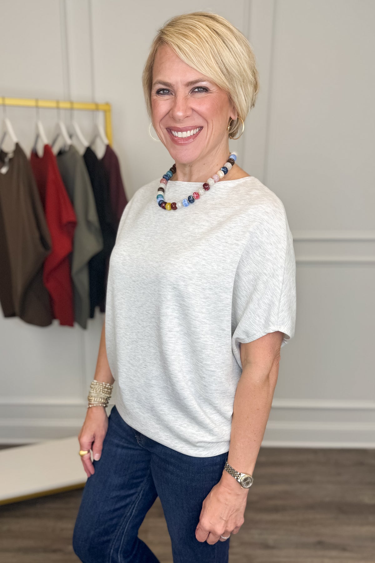 A smiling blonde woman wearing a light gray heathered short-sleeve top and dark blue jeans, standing in a boutique setting with a clothing rack visible in the background.