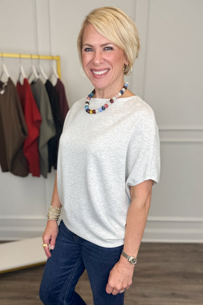 A smiling blonde woman wearing a light gray heathered short-sleeve top and dark blue jeans, standing in a boutique setting with a clothing rack visible in the background.