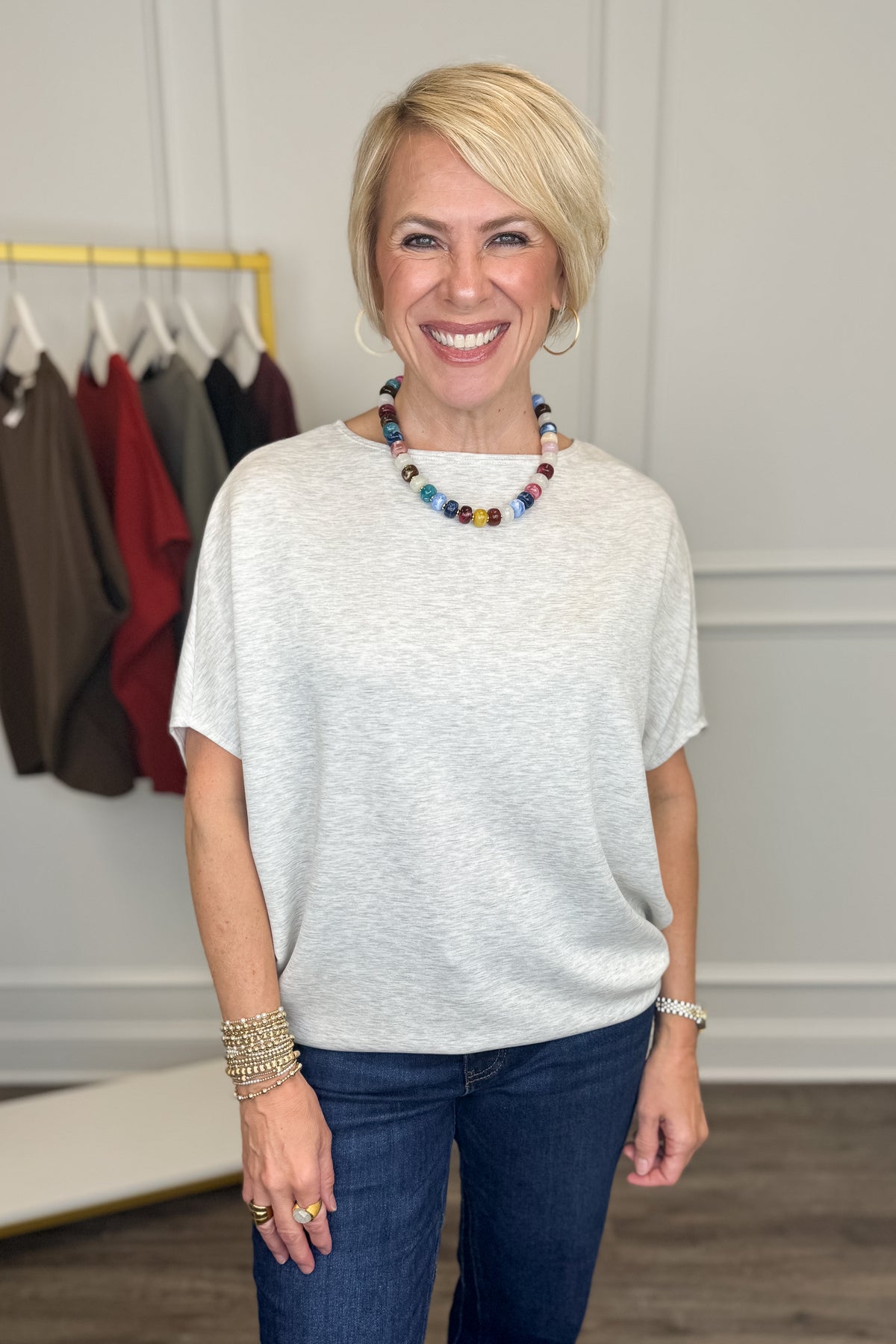 A smiling blonde woman wearing a light gray heathered short-sleeve top and dark blue jeans, accessorized with a colorful beaded necklace and bracelets, standing in a boutique setting.