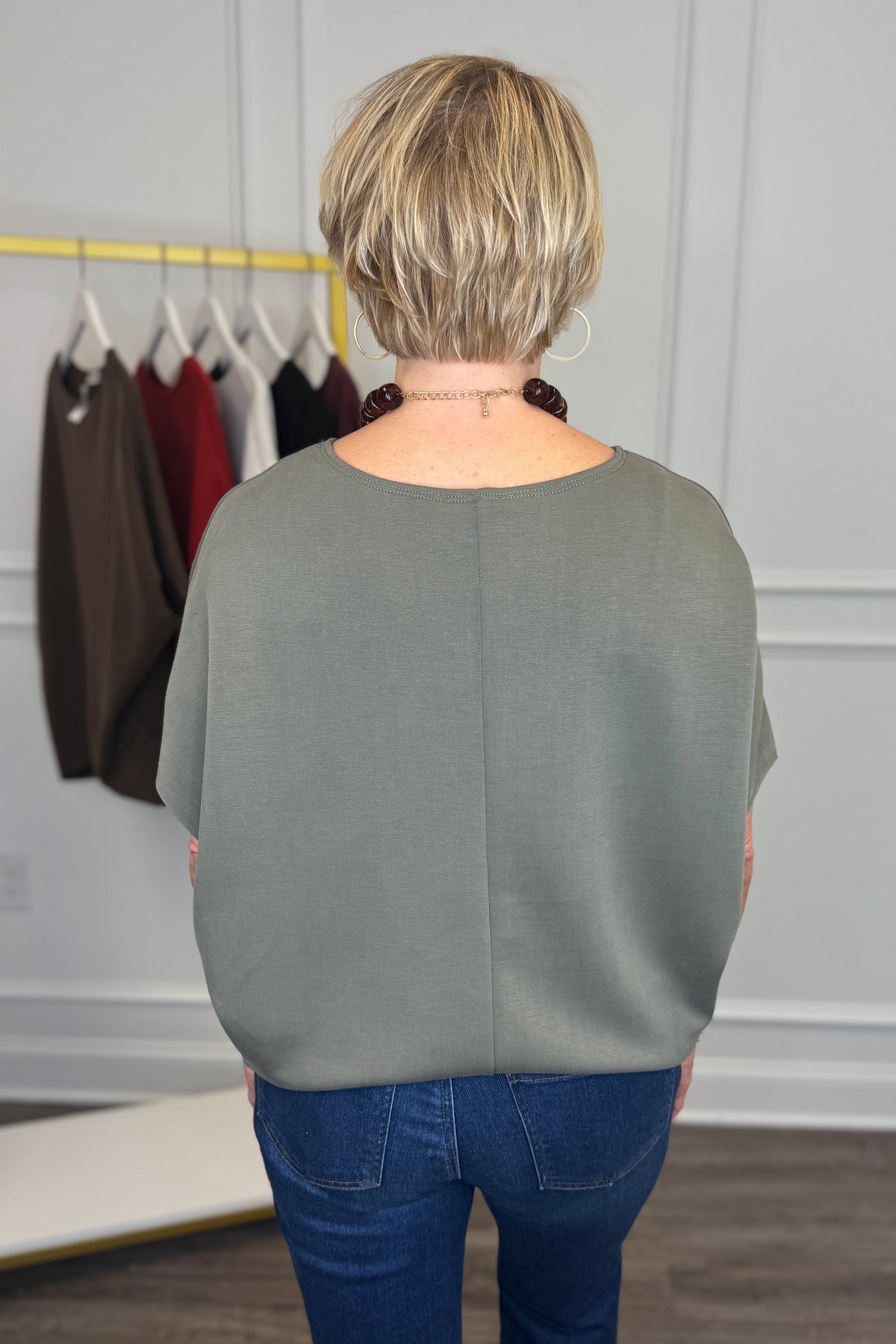 A woman viewed from behind wearing a muted sage green short-sleeve top and blue jeans, standing in a room with a clothing rack in the background.