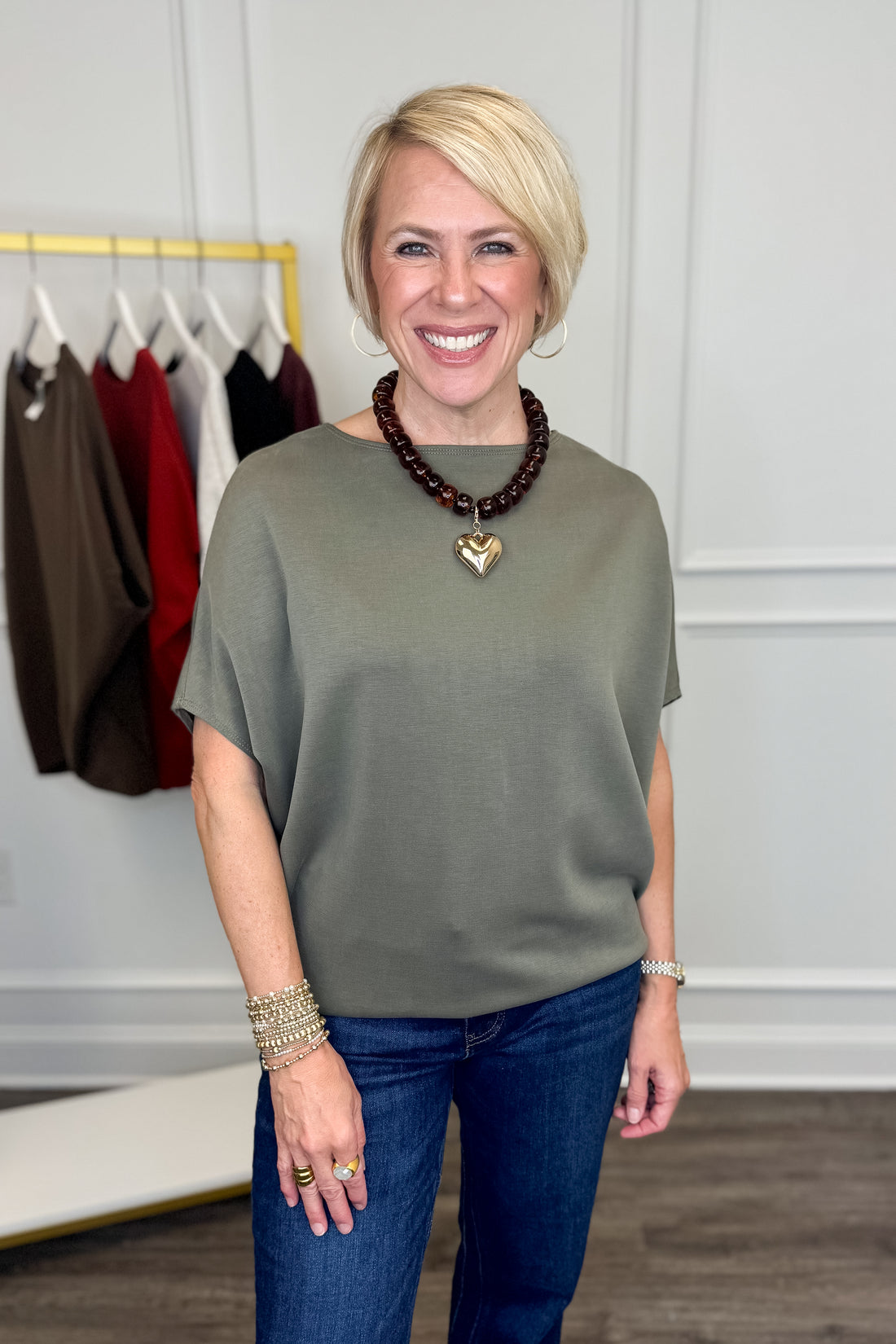 A smiling blonde woman wearing a short-sleeve olive green top and blue jeans, standing indoors in front of a clothing rack.