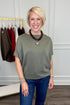 A smiling blonde woman wearing a short-sleeve olive green top and blue jeans, standing indoors in front of a clothing rack.