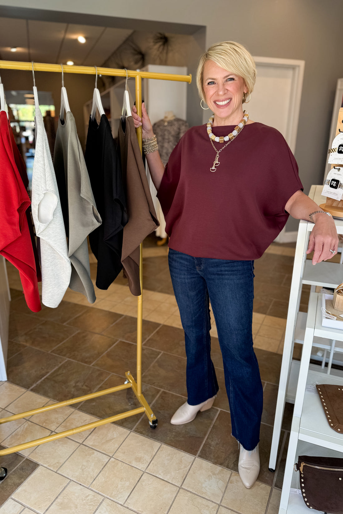A smiling blonde woman stands in a boutique next to a yellow clothing rack, wearing a burgundy short-sleeve top, dark blue jeans, and beige pointed-toe heels.