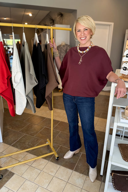A smiling blonde woman stands in a boutique next to a yellow clothing rack, wearing a burgundy short-sleeve top, dark blue jeans, and beige pointed-toe heels.