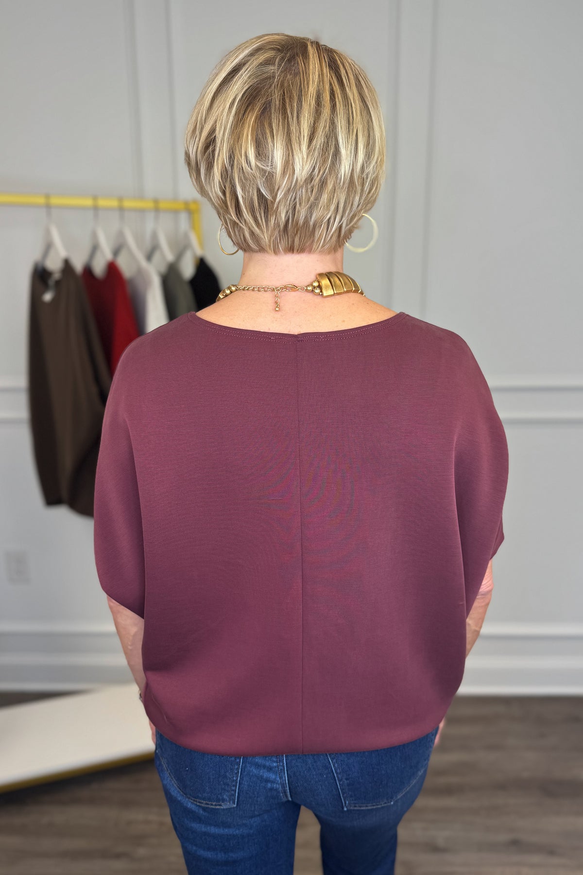 A woman viewed from behind wearing a mauve-purple short-sleeve top and blue jeans, standing in a boutique with a clothing rack visible in the background.