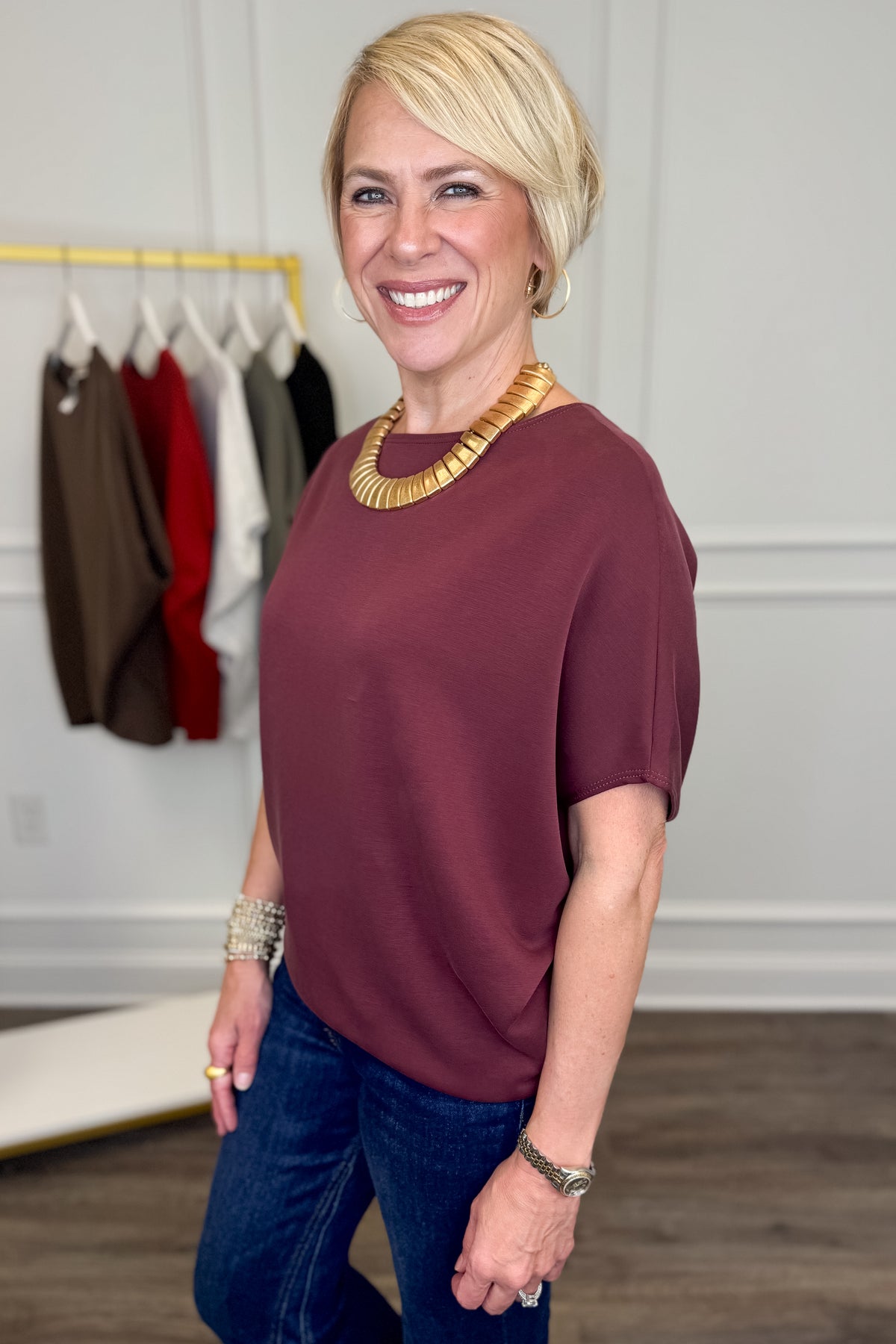 A smiling blonde woman wearing a short-sleeve scoop-neck top in burgundy and blue jeans, accessorized with a chunky gold necklace and bracelets, standing in a boutique setting.