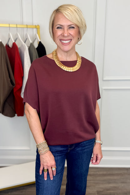 A smiling blonde woman wearing a short-sleeve, boat-neck burgundy top and dark blue jeans, accessorized with a chunky gold collar necklace and bracelets, standing in front of a white paneled wall with a clothing rack in the background.