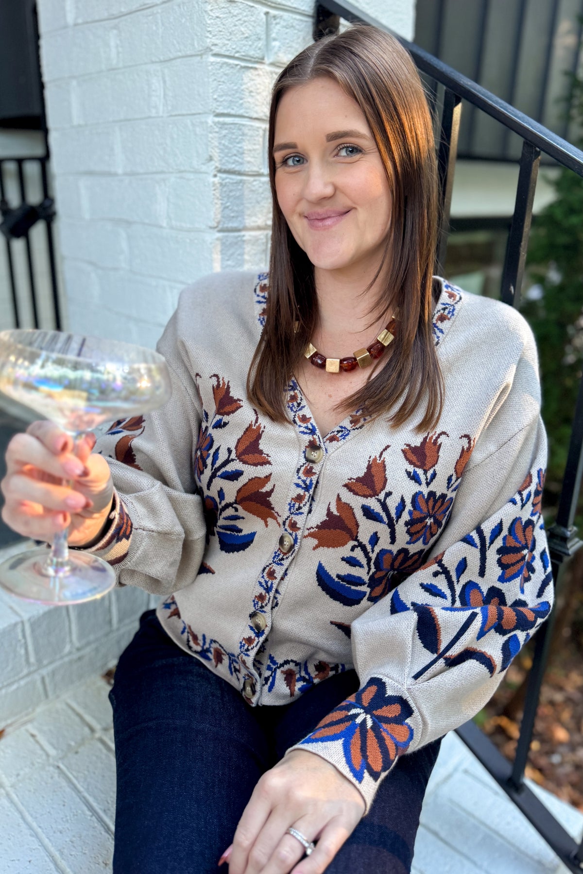 Front view of woman wearing Swagger Boutique’s Taupe Printed V-Neck Cardigan with jeans sitting outside at a party.