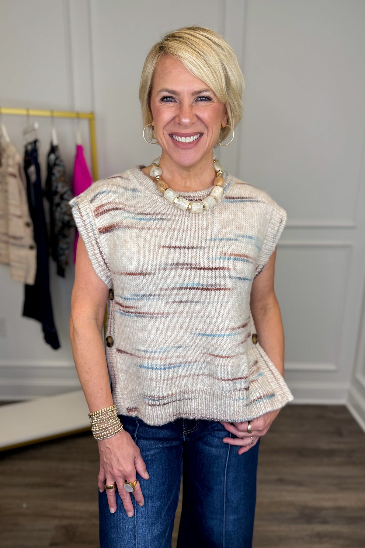 Front view of woman wearing Swagger Boutique’s Taupe, Brown &amp; Blue Button Detail Sleeveless Sweater with jeans with clothing hanging in the background.