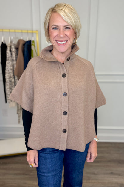 Front view of woman wearing Swagger Boutique’s Taupe Button Down Cape Jacket with jeans with clothing hanging in the background.