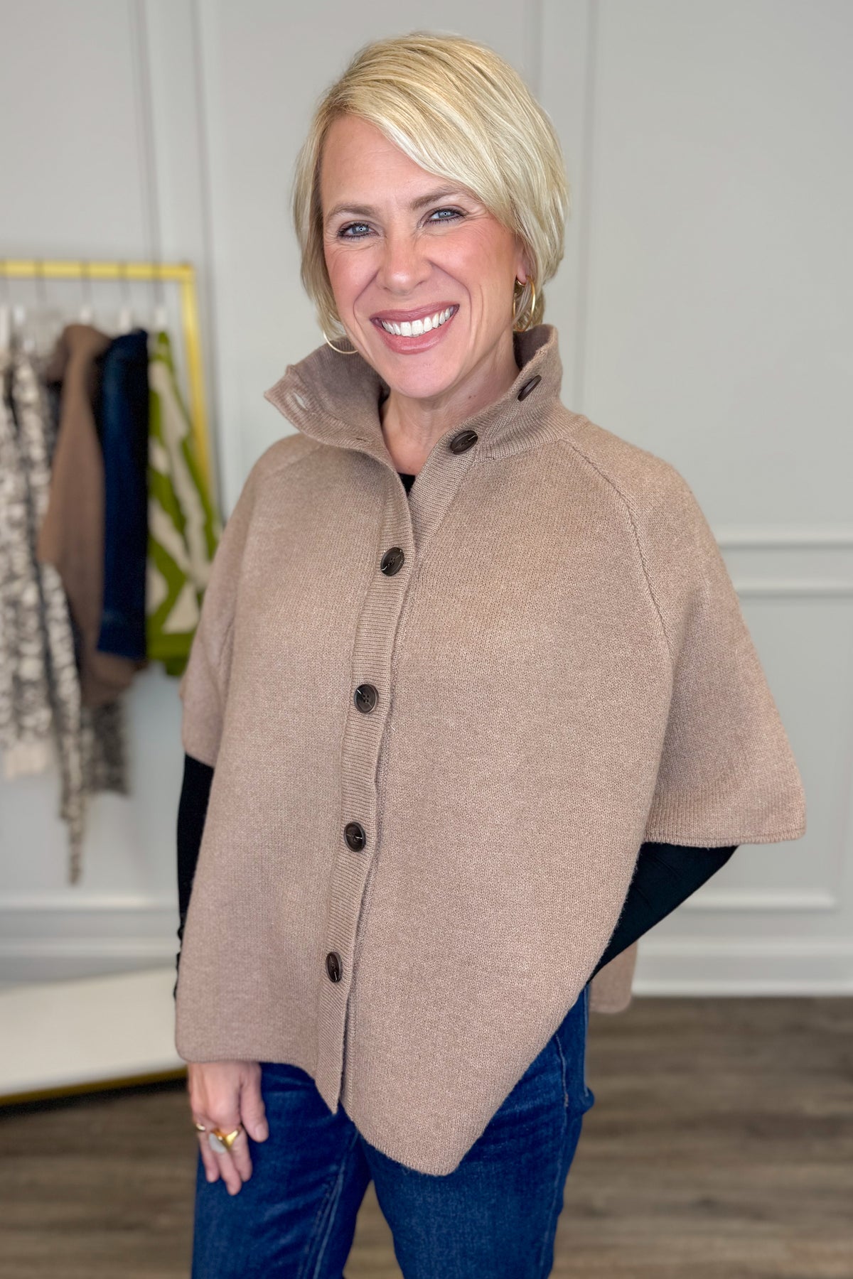 Side view of woman wearing Swagger Boutique’s Taupe Button Down Cape Jacket with jeans with clothing hanging in the background.