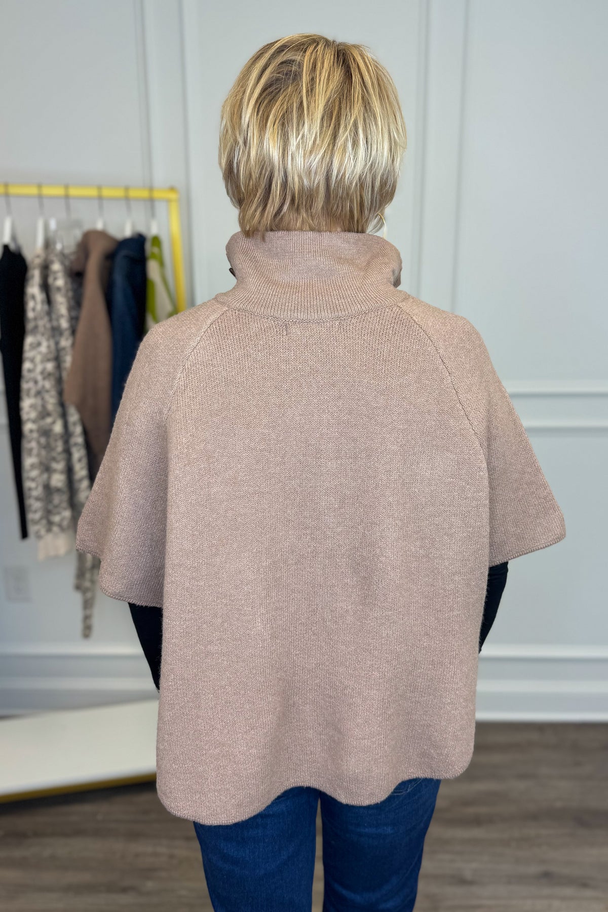 Back view of woman wearing Swagger Boutique’s Taupe Button Down Cape Jacket with jeans with clothing hanging in the background.