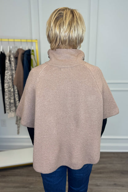 Back view of woman wearing Swagger Boutique’s Taupe Button Down Cape Jacket with jeans with clothing hanging in the background.