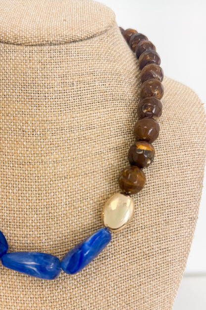 A close-up of a beaded necklace displayed on a beige fabric jewelry bust. The necklace features polished brown beads, a large smooth gold oval bead, and chunky blue beads.